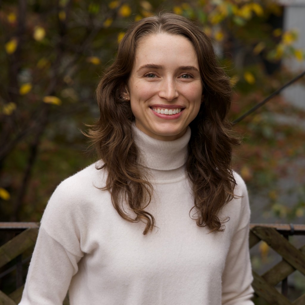 Shannon smiles warmly. She is a white woman with wavy brown hair, a big smile, green eyes, and a white sweater. Behind her is a tree with leaves just starting to turn yellow.